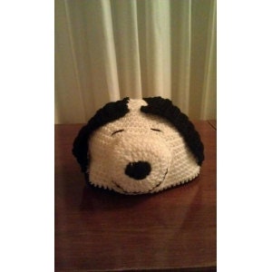 May include: A handmade crochet hat in the shape of a dog's head, featuring a white face with black accents for the ears and eyes. The hat is resting on a wooden surface, with a neutral-colored curtain in the background.
