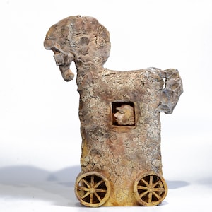May include: A textured, brown ceramic sculpture of a horse-shaped figure on wheels. The body features a rectangular cutout containing a small, sculpted human head in profile. The surface has a cracked, weathered appearance with earthy tones.