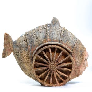 May include: A textured ceramic sculpture shaped like a fish with a weathered, mottled brown and gray finish. The body features a prominent circular wheel with spokes on the side, rivet-like details along the edges, and a human-like face profile.