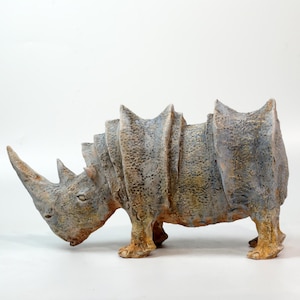 May include: A ceramic rhinoceros sculpture featuring a textured, mottled surface in shades of dusty blue, grey, and earthy orange. The figure displays prominent, layered armour-like plates along its back and a single large horn on its snout.