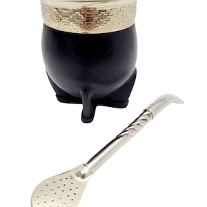 Peut inclure: Un récipient à maté noir avec un bord doré et une bombilla argentée. Le récipient a une forme arrondie et repose sur trois petits pieds. La bombilla a une extrémité en forme de cuillère avec de petits trous.