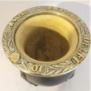 May include: A traditional wooden mate gourd with a brass rim. The rim is engraved with the words "RENACA DEL RIO 10" and a floral design.