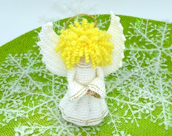 May include: A crocheted angel ornament with white wings, a yellow halo, and a white dress. The angel is sitting on a green surface with white snowflake decorations.
