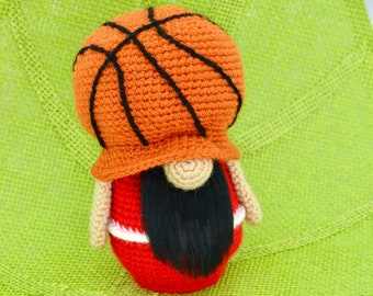 May include: A crocheted gnome wearing a basketball-shaped hat and a red and white jersey. The gnome is sitting on a green surface.