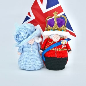 Può includere: Una bambola all'uncinetto della regina con un vestito e un cappello blu con un fiore, in piedi accanto a una bambola all'uncinetto del re con un'uniforme rosso e oro e una corona viola. Una piccola bandiera dell'Unione Jack è dietro di loro.