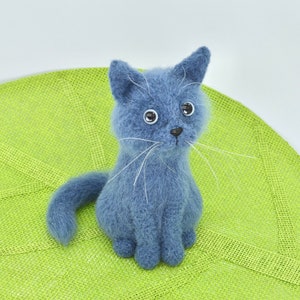 May include: A blue felted cat toy with white whiskers sitting on a green mat.