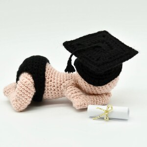 May include: A crocheted baby doll wearing a black graduation cap and lying on a white surface with a rolled up diploma tied with a gold ribbon.