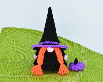 May include: A crocheted witch gnome wearing a black pointed hat with a purple brim and orange hair. The gnome is holding a small purple crocheted pumpkin.