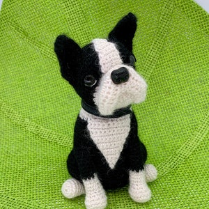 May include: A black and white crocheted Boston Terrier dog sitting on a green woven mat. The dog has black ears, a black nose, and white fur on its chest and paws.