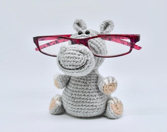 May include: A gray crocheted hippopotamus wearing pink glasses. The hippopotamus is sitting on a white surface.