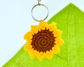 Puede incluir: Un llavero de girasol tejido a crochet con un centro marrón y pétalos amarillos. El llavero está unido a un anillo de llave plateado.