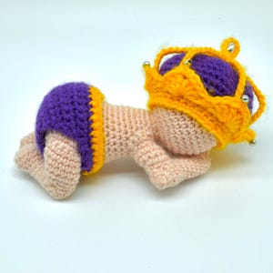 Op de afbeelding: Een gehaakte babypop met een paarse en gele luier en een gele kroon met zilveren accenten.