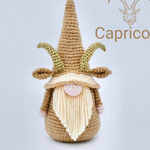 May include: Crocheted Capricorn gnome with a brown hat, gold horns, and a cream-colored beard. The gnome is standing on a white background. The text "Capricorn" is written in brown above the gnome.