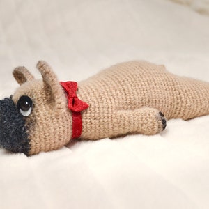 May include: A crocheted stuffed dog toy in a light brown colour with a red bow tie. The dog is lying on its side with its head turned to the side.