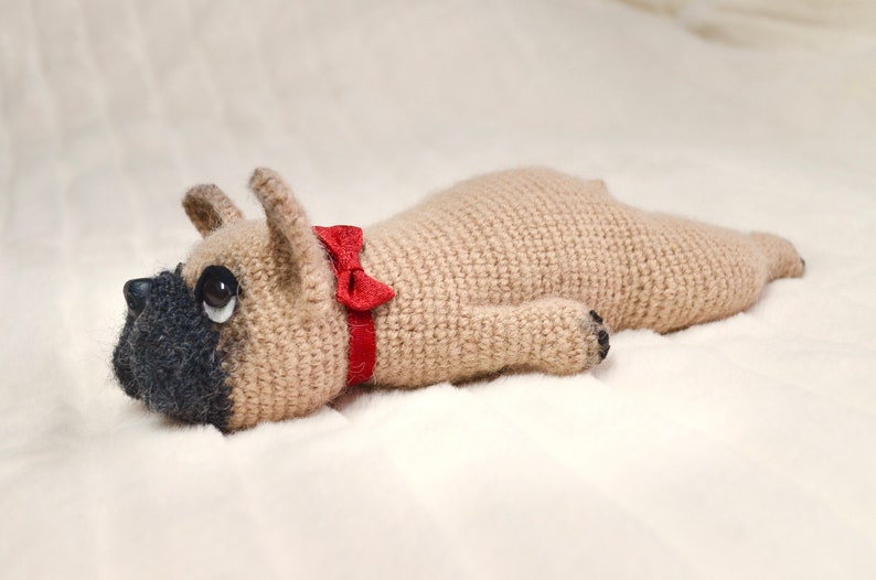 May include: A crocheted stuffed dog toy in a light brown colour with a red bow tie. The dog is lying on its side with its head turned to the side.