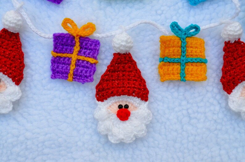 May include: A crocheted garland with Santa Claus and gift box ornaments. The garland is white and the ornaments are red, white, purple, yellow, and blue.