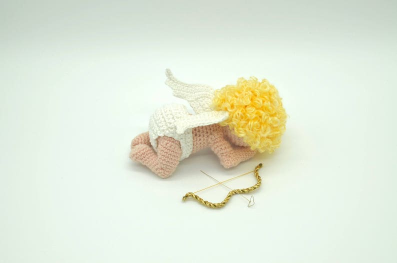 May include: A crocheted amigurumi cupid doll with a white diaper, pink skin, and yellow curly hair. The doll is lying on its side with its wings spread out behind it. A gold bow and arrow is lying next to the doll.