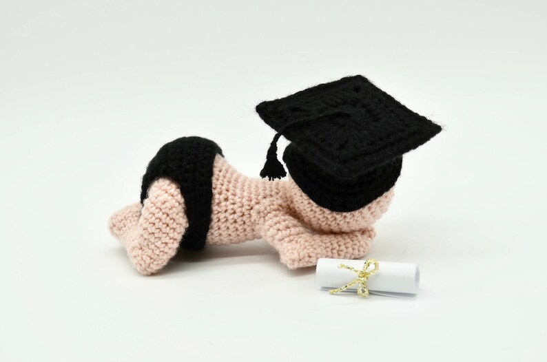 May include: A crocheted baby doll wearing a black graduation cap and lying on a white surface with a rolled up diploma tied with a gold ribbon.