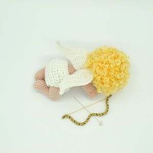May include: A crocheted angel figurine with a white dress, pink skin, and yellow curly hair. The angel is lying on its side with its wings spread out. A gold bow and arrow is lying next to the angel.