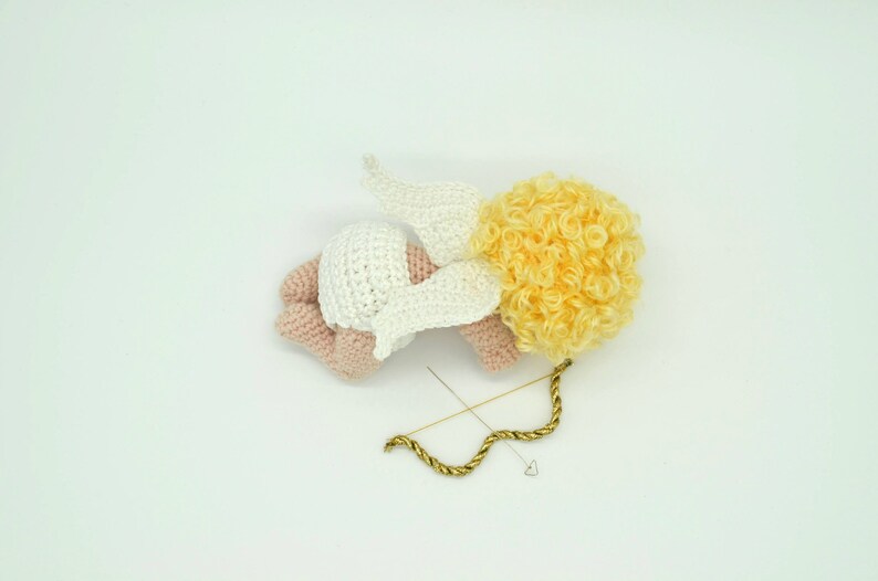 May include: A crocheted angel figurine with a white dress, pink skin, and yellow curly hair. The angel is lying on its side with its wings spread out. A gold bow and arrow is lying next to the angel.