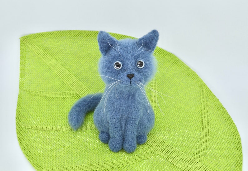 May include: A blue knitted cat toy sitting on a green leaf-shaped mat. The cat has white whiskers and black eyes.
