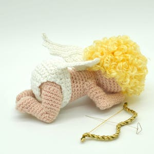 May include: A crocheted amigurumi cupid doll with a white diaper, white wings, and yellow curly hair. The doll is lying on its side with a gold bow and arrow beside it.