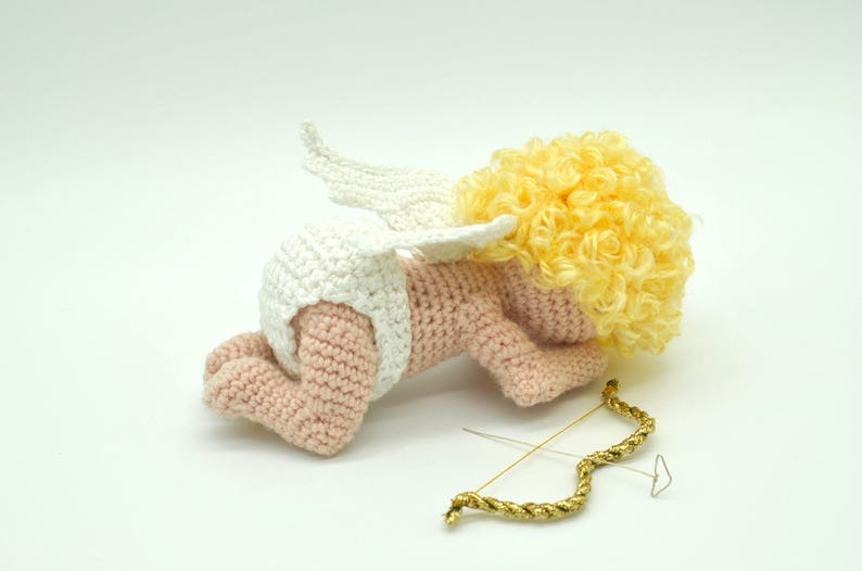 May include: A crocheted amigurumi cupid doll with a white diaper, white wings, and yellow curly hair. The doll is lying on its side with a gold bow and arrow beside it.