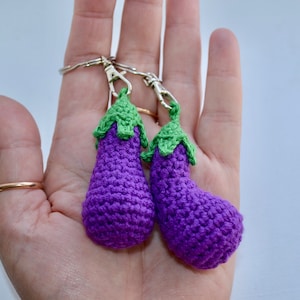 Puede incluir: Dos llaveros de berenjena de ganchillo morado con tapas verdes y anillos de llaves plateados.