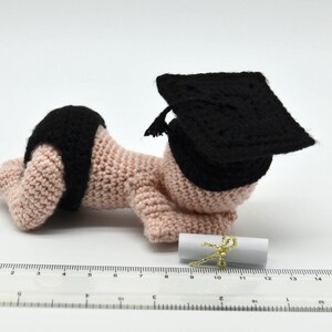 May include: A crocheted baby doll wearing a black graduation cap and holding a rolled up diploma. The doll is lying on its stomach with its arms outstretched.