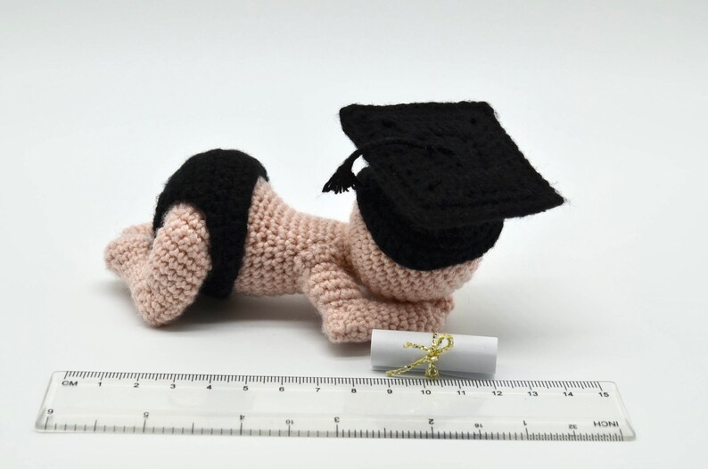 May include: A crocheted baby doll wearing a black graduation cap and holding a rolled up diploma. The doll is lying on its stomach with its arms outstretched.