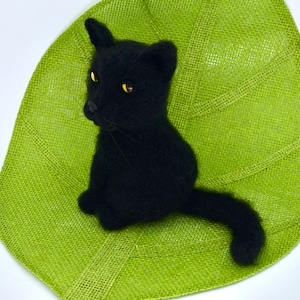 May include: A black crocheted cat sitting on a green leaf-shaped placemat. The cat has yellow eyes and a long tail.