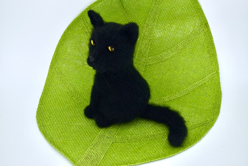 May include: A black crocheted cat sitting on a green leaf-shaped placemat. The cat has yellow eyes and a long tail.