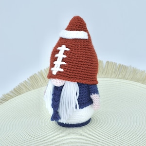 May include: A crocheted gnome wearing a brown and white football hat, a blue sweater, and a white beard. The gnome is sitting on a white round placemat with a fringe.