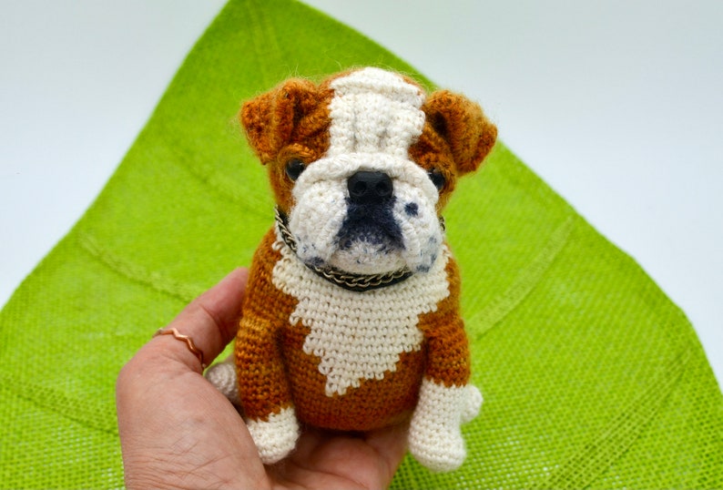 May include: A crocheted bulldog plush toy with brown and white fur, wearing a black collar. The toy is sitting on a green surface.