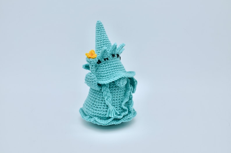 Puede incluir: Un gnomo de crochet con un sombrero puntiagudo, una flor amarilla y una barba trenzada. El gnomo es azul claro y tiene ojos negros.