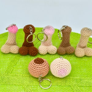 May include: A set of seven crocheted keychains in various colors, including brown, pink, and beige. The keychains are shaped like anatomical body parts. The keychains are displayed on a green surface.