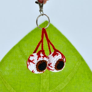 Puede incluir: Un llavero de ganchillo con dos ojos blancos de ganchillo con venas rojas y pupilas marrones. El llavero está unido a un llavero plateado.