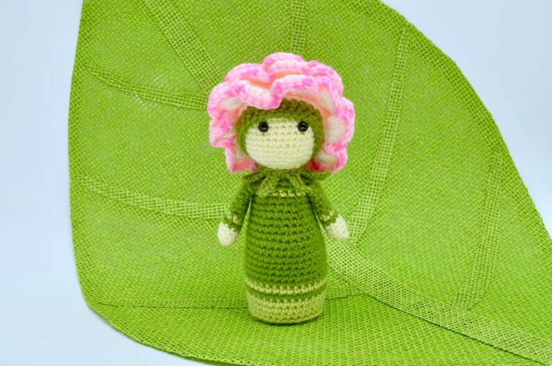 Op de afbeelding: Een gehaakte pop met een roze bloemenhoed en een groene jurk. De pop zit op een groen gehaakt blad.