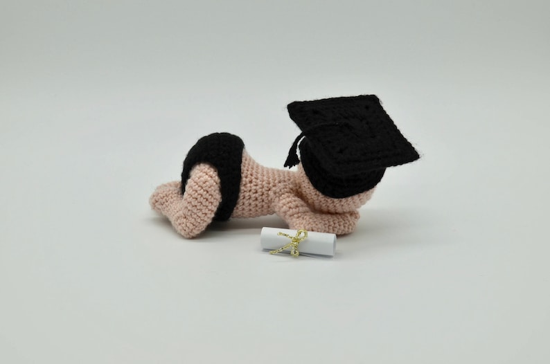 May include: A crocheted baby doll wearing a black graduation cap and a diaper, holding a rolled up diploma.