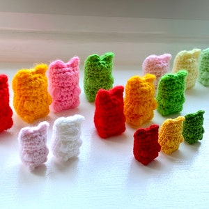 May include: A selection of small, handmade crochet figures in a range of colours including red, yellow, pink, green, and white. The figures have a bear-like shape with small ears, and are arranged on a white surface.