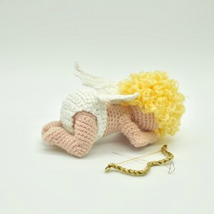 May include: A crocheted amigurumi cupid doll with a white diaper, pink skin, and yellow curly hair. The cupid is lying on its stomach with its wings spread out behind it. A gold bow and arrow is lying on the white surface.
