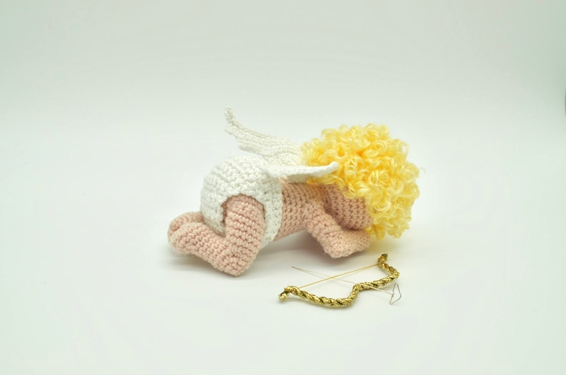 May include: A crocheted amigurumi cupid doll with a white diaper, pink skin, and yellow curly hair. The cupid is lying on its stomach with its wings spread out behind it. A gold bow and arrow is lying on the white surface.