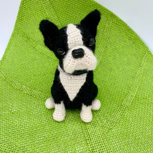 Pu&ograve; includere: Un giocattolo per cani Boston Terrier all'uncinetto, nero e bianco con una macchia bianca sul petto, seduto su un tappetino verde intrecciato.