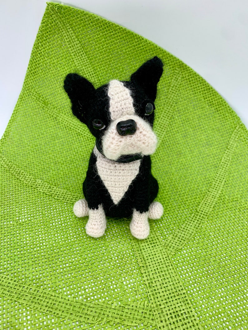 Pu&ograve; includere: Un giocattolo per cani Boston Terrier all'uncinetto, nero e bianco con una macchia bianca sul petto, seduto su un tappetino verde intrecciato.