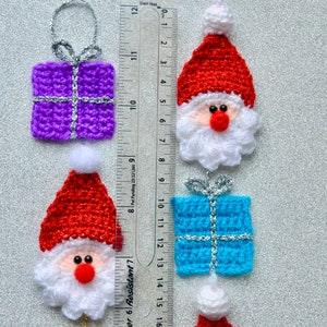 May include: Five crocheted Christmas ornaments, including two Santa Claus figures with red hats and white beards, two gift boxes, one purple and one blue, and one Santa Claus hat.