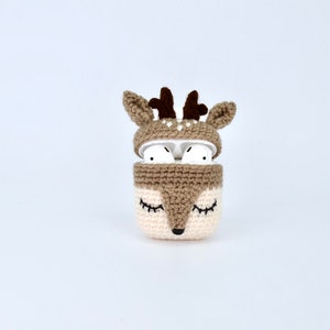 May include: A crocheted AirPods case in the shape of a deer. The case is primarily beige and brown, with a deer face design on the front and antlers on top. The AirPods are visible inside the open case.