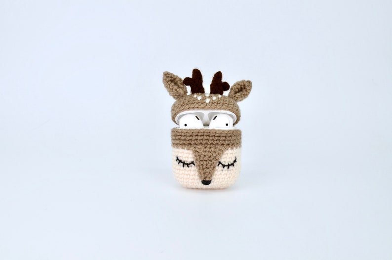 May include: A crocheted AirPods case in the shape of a deer. The case is primarily beige and brown, with a deer face design on the front and antlers on top. The AirPods are visible inside the open case.