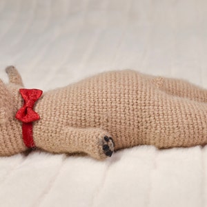 May include: A crocheted beige pug dog toy wearing a red bow tie. The toy is lying on a white fluffy surface.