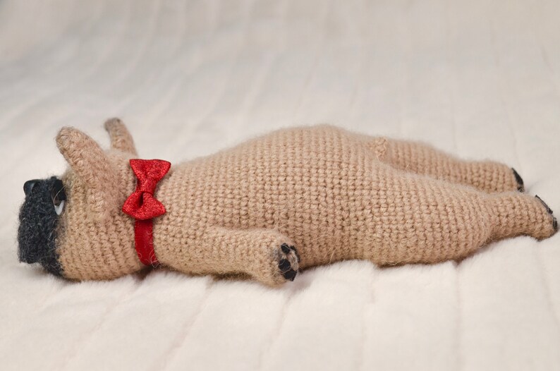 May include: A crocheted beige pug dog toy wearing a red bow tie. The toy is lying on a white fluffy surface.
