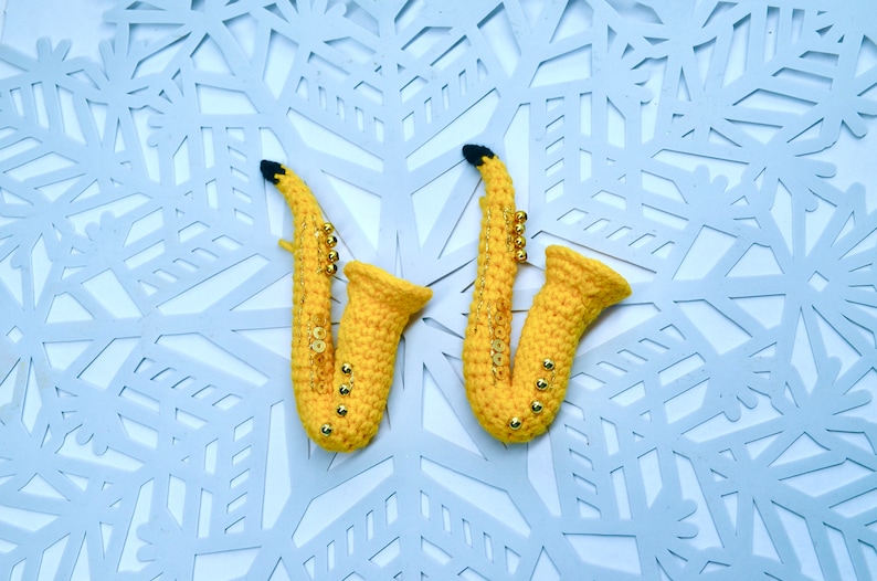May include: Two yellow crocheted saxophones with gold beads on a white snowflake background.